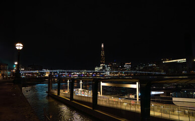 Fototapeta na wymiar Uber boat dock and walkway, public transportation service along the River Thames, at night with the Millennium Bridge, streetlights and the Shard skyscraper illuminated by the river water.