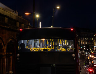 View from the rear of passengers or commuters inside a London double decker bus traveling at night along a central London street with motorbikes and cyclists traveling alongside. - obrazy, fototapety, plakaty