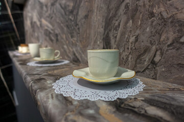 A white cup with a saucer and a lace napkin stands on a marble window sill.