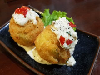 A close-up view of a plate containing two deep-fried balls. The balls are golden brown and round, with sesame seeds sprinkled on their surface. A thick orange sauce is drizzled over the balls.