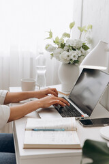 Close-up view of woman hands typing text on a laptop keyboard on white table with bottle of drinking water and flowers in vase on the background. Concept of freelancing, remote work, online learning