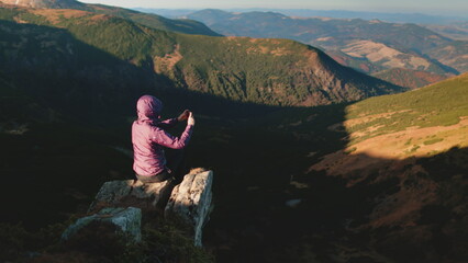 Trekking person in autumn Carpathian mountains sit on rock top peak make panoramic shot of sunset canyon hill on mobile phone. Outdoor lifestyle travel trekking hiking holiday vacation.