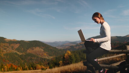 Young woman sitting on a rustic fence, typing on laptop and enjoying autumn sunset mountains nature. Work outdoor, online digital nomad remote job in Carpathian mountains. Outdoor lifestyle travel