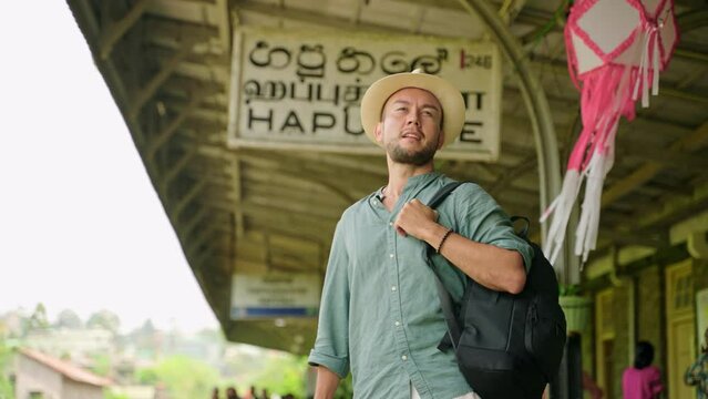 Asian tourist with backpack navigates Haputale railway station. Man with camera explores authentic Sri Lankan transport hub. Traveler in hat ready for train journey through lush highlands. Slowmo