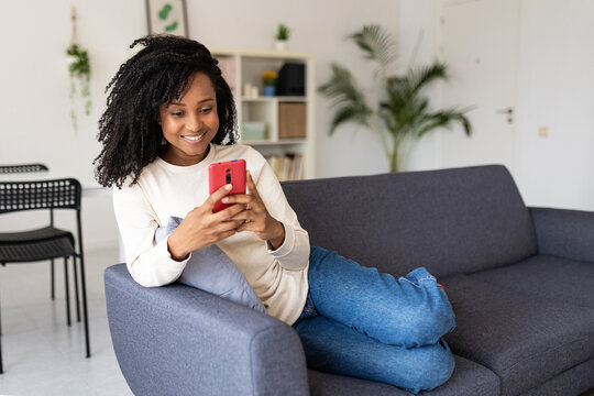 Happy young african american woman holding mobile phone device while relaxing on sofa at home.