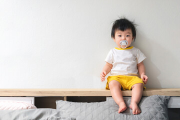 Asian baby with pacifier in her mouth is sitting on the bed in bedroom.