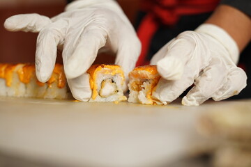 Japanese sushi is cutted and plated by gloves wearing hand chef with sharp knife, shrimp and sauces are visible in between sticky rice and bamboo role and firing the sushi role with burner 