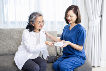 A senior asian woman sits on sofa getting her blood pressure checked by a home visiting nurse,older adult female looking at pressure gauge and asking about the examination results