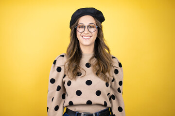 Young beautiful brunette woman wearing french beret and glasses over yellow background with a happy face standing and smiling with a confident smile showing teeth