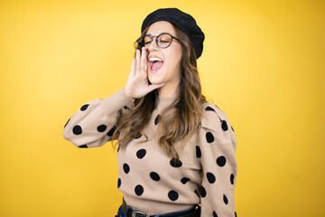 Young beautiful brunette woman wearing french beret and glasses over yellow background shouting and...
