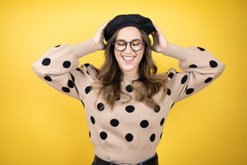 Young beautiful brunette woman wearing french beret and glasses over yellow background suffering...