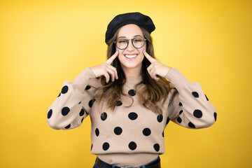 Young beautiful brunette woman wearing french beret and glasses over yellow background very happy and excited making winner gesture with raised arms, smiling and screaming for success.
