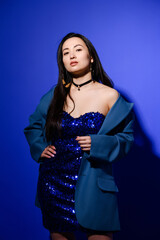 Stylish young asian woman posing in dress with sequins and jacket on blue background 