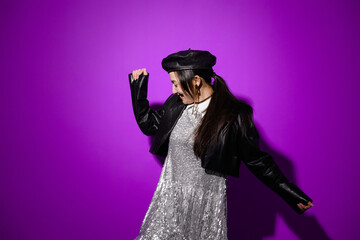 Cheerful asian woman in leather beret and dress dancing on purple 
