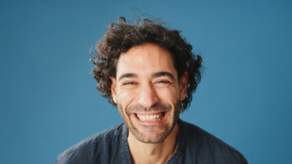 Close up of laughing attractive man with curly hair, dressed in blue shirt, crossing his arms and...