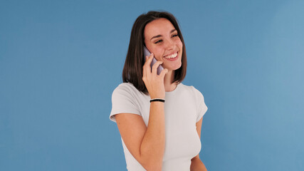 Laughing beautiful young brunette woman dressed in white top talking on smartphone isolated on blue...