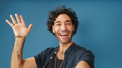 Smiling man with curly hair, wearing blue shirt, looking at camera making video call, welcome sign...