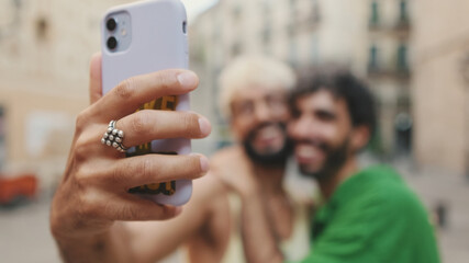 Close-up of hand, gay couple stands hugging and takes selfie on mobile phone, background blurre