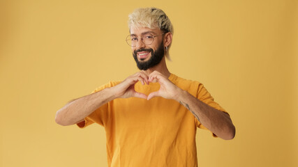 Guy with glasses, dressed in yellow T-shirt, showing representing heart in shape of fingers gesture...
