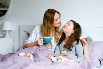 Obraz na płótnie Canvas Lovely young mother and cute school kid girl cuddling together in bed in morning. Happy family of young single woman and beautiful daughter having breakfast, eating on mother's day.