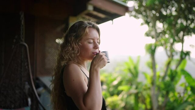 Relaxed woman sips tea on resort terrace, mountain backdrop, serene morning vibe. Curly-haired lady enjoys hot beverage, daybreak freshness, wellbeing moment. Luxury escape, tranquility. Slow motion.