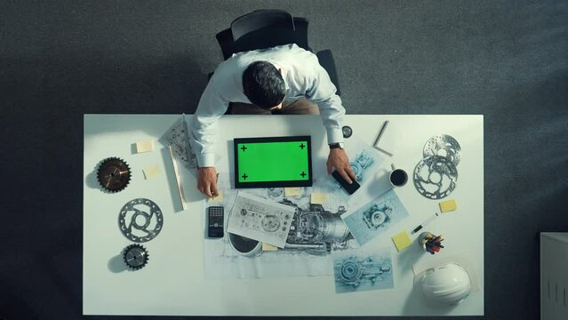 Top view of engineer looking at phone while working on tablet with green screen at meeting table with turbine engine or jet engine sketch design and various metal gear scatter around. Alimentation.