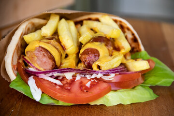 Flavorful tortilla wrap is a delightful fusion of savory sausages, crispy French fries, fresh green salad, juicy tomatoes, zesty onions, and shredded cabbage, all wrapped up with tangy mustard