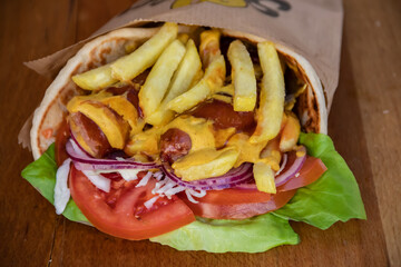Flavorful tortilla wrap is a delightful fusion of savory sausages, crispy French fries, fresh green salad, juicy tomatoes, zesty onions, and shredded cabbage, all wrapped up with tangy mustard