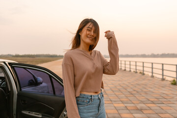 Photo of asian woman smiling and standing next to the car near the river at sunset time, chilling traveling on vocation