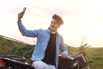 Curly haired Caucasian young man sitting on the side of his motorbike with raised hand taking a...
