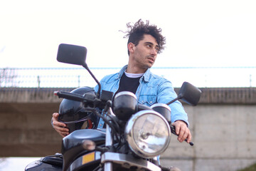 Portrait of curly haired Caucasian young man holding his motorcycle and helmet, looking to the side with bridge in the background. Lifestyle concept
