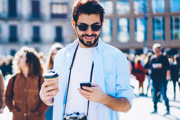 Cheerful bearded man in stylish sunglasses holding coffee to go while reading incoming sms message...