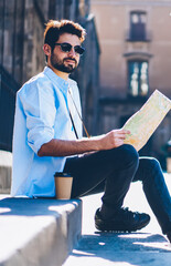 Bearded tourist in black sunglasses sitting with travel map in architectural city during trip in...