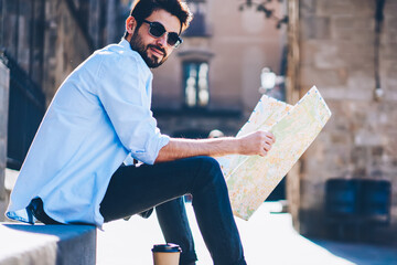 Portrait of cheerful bearded tourist in sunglasses sitting outdiirs on street of architectural city with old constructions while holding travel map for searching destination of showplaces