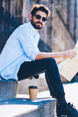 Portrait of handsome casual dressed traveler in stylish sunglasses enjoying summer vacation in old town while sitting with map in hands on street.Positive male tourist during trip in sunny day