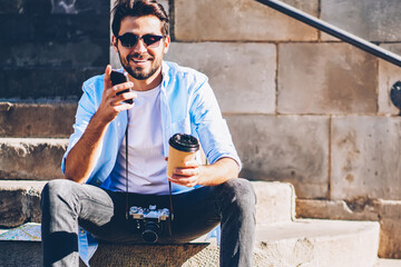 Cheerful bearded man with vintage camera dialing number on phone while sitting on stairs of...