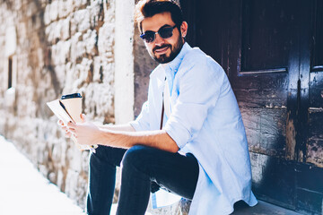 Portrait of handsome bearded toursit in sunglasse holding map and coffee in hand while resting on...