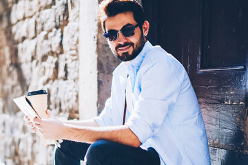 Portrait of handsome bearded toursit in sunglasse holding map and coffee in hand while resting on...