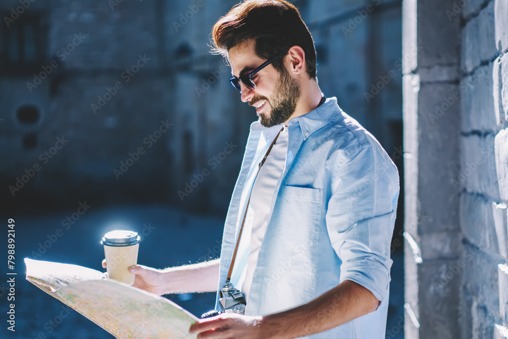 Wall mural Happy stylish tourist in black sunglasses and casual outfit holding coffee takeaway in hand and searching right route to location of showplaces on travel map strolling in urban setting during trip