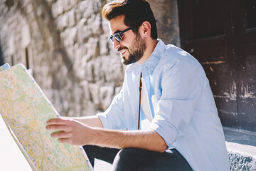 Smiling bearded traveler in spectacles reading map to find right direction of showplaces sitting outdoors on street.Positive young man tourist in sunglasses search destination during trip in old town