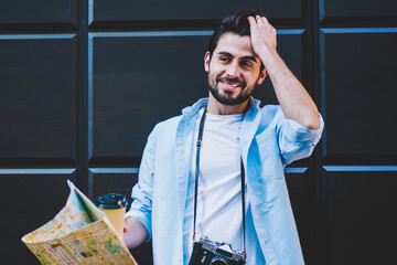 Cheerful bearded male tourist touching hair on black promotional background with copy space area.Handsome traveler with vintage camera holding map in hand while standing outdoors on street