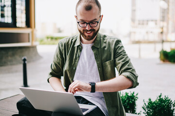 Caucasian young man looking at smartwatch while working with laptop computer outdoors, hipster guy...