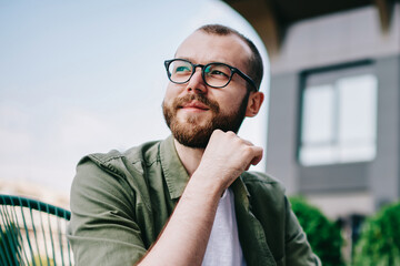 Young handsome male 20 years old in eyeglasses sitting at street cafe during free time with new modern laptop, bearded man freelancer  thoughtfully looking aside and thinking about distance work