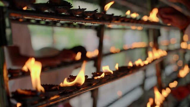 Flames flicker on oil lamps at Buddhist temple, devotees perform rituals with reverence in serene setting. Worshipers lamps, offering prayers, practicing meditation, spiritual homage in sacred shrine.