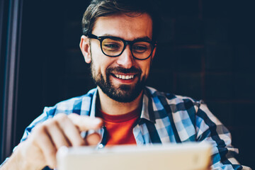 Positive bearded hipster guy in eyeglasses watching broadcasting on modern tablet connected to 4G...