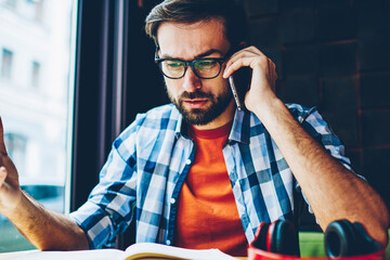 Angry bearded young man in eyeglasses communicating on smartphone device sitting at desktop with...