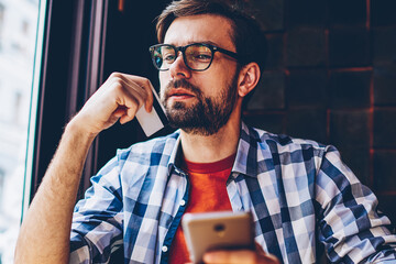 Thoughtful bearded hipster guy with business card in hands thinking on working proposal sitting in cafe with smartphone.Pensive young man pondering on money transaction via 4G internet on cellular