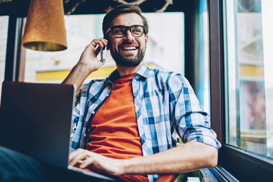 Cheerful bearded hipster guy calling on smartphone and laughing during mobile conversation looking out of window.Positive male blogger talking with friend on telephone while working at netbook - Powered by Adobe