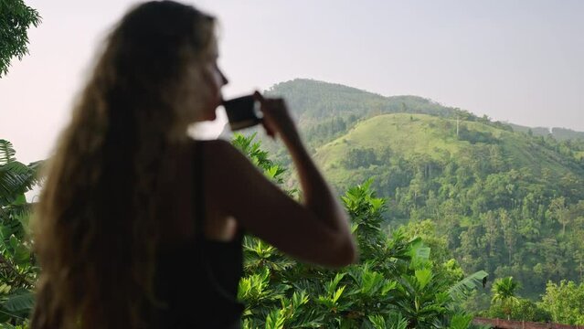 Elegant woman enjoys hot beverage on upscale resort patio overlooking Ella Rock, rich scenery of Sri Lanka. Relaxing, sips coffee, tranquil mountain view surround, travel luxury experience. Slowmo