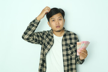 Young Asian man wearing brown flannel shirt holding paper money and showing confused face expression